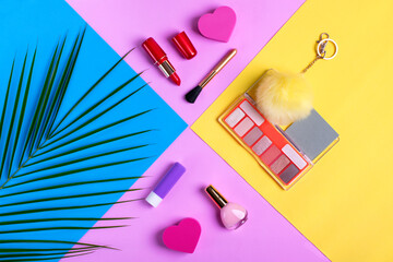 Makeup products on colorful geometric background. Eyeshadow palette, red lipstick, lip balm, nail polish, makeup brush, yellow pompon and heart-shaped sponge. Cosmetic flat lay