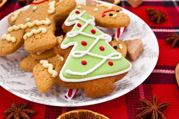 Christmas greeting card. Fir shaped cookie with gingerbread men in a plate, sugar hearts, dried orange slices, red berries and spices on a red fabric background.