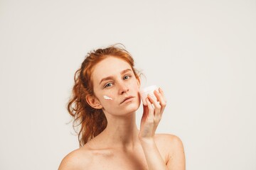 Portrait of natural redhead young woman with long wavy hair and freckles, holds nutrition face cream. Isolated over white background. Natural beauty and health
