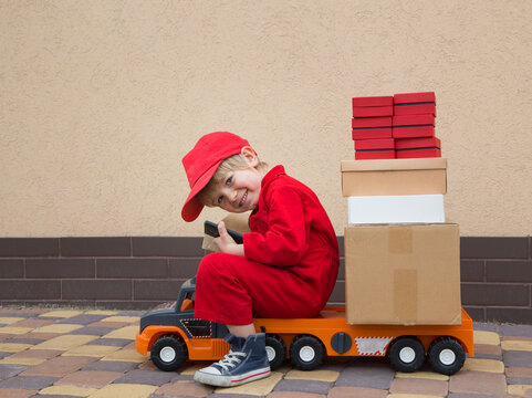 happy toddler boy in a red overalls and a cap sits on a large toy car loaded with parcels - boxes. games for kids - postman, be like dad