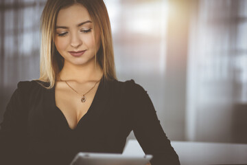 Successful young businesswoman is using a computer tablet, while working in a sunny cabinet of her firm. Business headshot or portrait of a secretary, sitting at the desk in an office