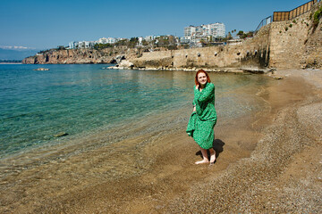 Early spring in turkish antalya, young white woman in green evening dress walks barefoot along beach.