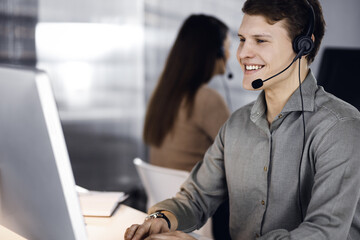 Young dark-haired guy in a green shirt and headsets is talking to a client, while sitting at the...