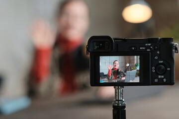 Portrait of young woman waving at camera while sitting at the table with laptop and recording herself on camera