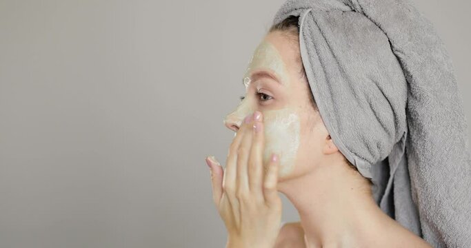 Side view of beautiful young blue-eyed girl applying clay face mask after taking a bath. Facial beauty and skin care concept in home spa treatments