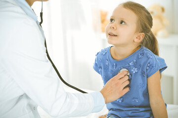 Doctor examining a child by stethoscope. Happy smiling girl patient dressed in blue dress is at usual medical inspection. Medicine concept