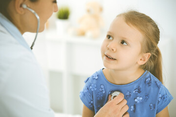 Fototapeta premium Doctor examining a child by stethoscope. Happy smiling girl patient dressed in blue dress is at usual medical inspection. Medicine concept
