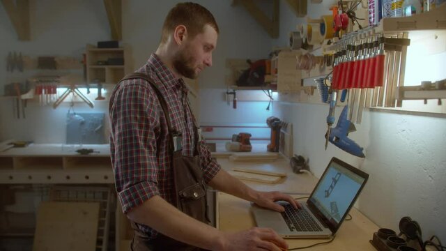 young Caucasian carpenter with beard measure the boards on the table and using laptop for checking