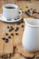 Top view of white jug of milk and white cup of coffee, on rustic wooden table with coffee beans, selective focus, vertical