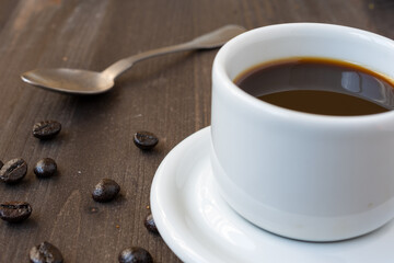 Close-up of white cup with coffee and spoon, on wooden table with coffee beans, selective focus, horizontal