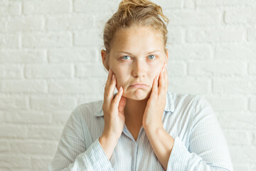 Close up serious upset woman touching her face with two hands while looking at the camera. Captured in studio with light gray background