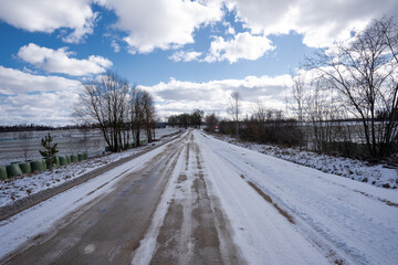 Obraz premium rural dirt road in spring with white snow and ice, shrubs growing along the edges and lots of fluffy clouds in the beautiful blue sky