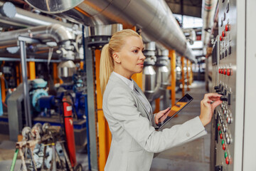 Female blond supervisor standing in heating plant next to dashboard, adjusting settings and holding tablet.