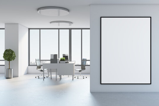 Blank white poster in black picture frame on light wall in stylish open space office with modern furniture, tree in metallic flowerpot on concrete floor and city view from big window