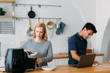 Beautiful young caucasian wife in kitchen cooking healthy oil free meal in modern air fryer with handsome husband working on laptop in kitchen at home
