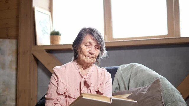 Senior woman reads a book in a comfortable chair. Reading books, new knowledge in old age, scientific literature