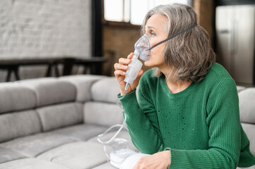 Upset senior woman feeling unwell, has shortness of breath, asthma, using oxygen mask sitting on the couch at home. Sick mature woman makes inhalation with a nebulizer at home, treating herself - obrazy, fototapety, plakaty
