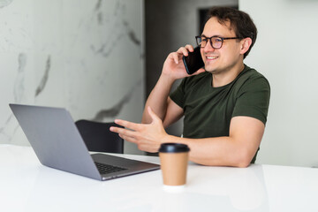 Optimistic man speaking on mobile phone and using laptop at kitchen