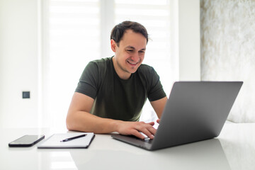 Fototapeta premium Handsome young man calculating domestic bills or taxes, household expenses, managing budget, paperwork, checking financial documents, using laptop in modern kitchen