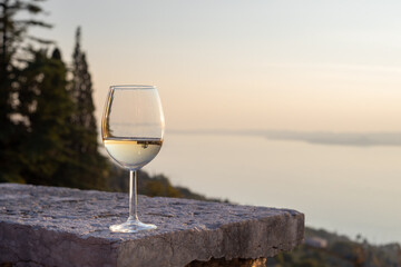 a glass of white wine stands on a terrace wall and in the background you can see a lake and mountains. 
