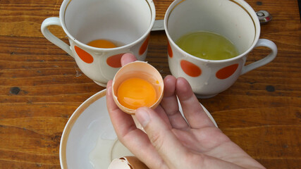 Raw egg yolk separated inside half of broken eggshell holding in female fingers. Preparation of food ingredient for cooking ang baking. Retro kitchen crockery on background