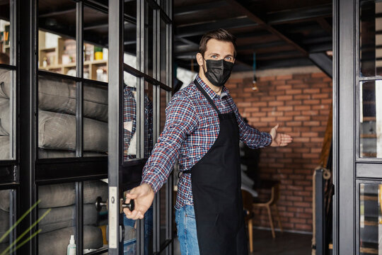 Welcome to the restaurant, we are open. A male waiter in a plaid shirt and with a black apron wears a protective face mask and greets guests at the entrance to the facility and opens the door for them