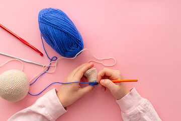 Children's hands in the process of crocheting toys from blue and beige yarn. Pink background....