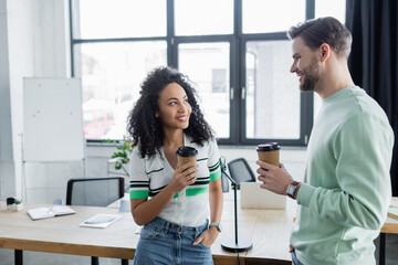 cheerful multicultural managers holding coffee to go while talking in office