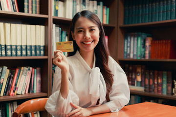 Young happy Asian business woman holding credit card and smart phone with book shlf full of books in the vintage style reading room.