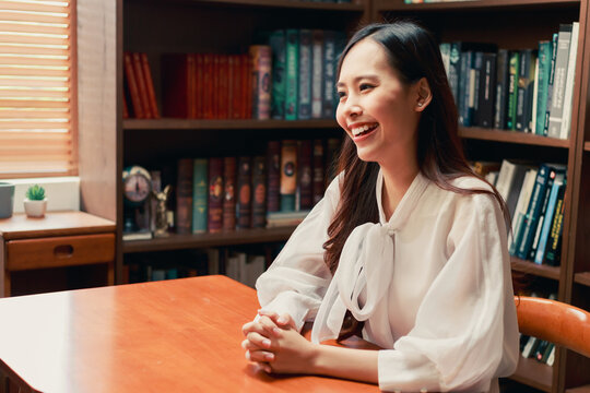 Happy Asian young knowledgable business woman sitting in library with various book with books on the bookshelf background