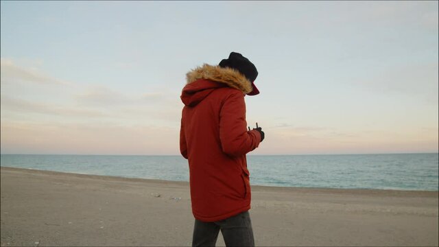 Ragazzo con cappotto arancione sulla spiaggia pilota un drone visto di spalle