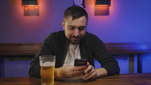 Portrait of man with glas of beer looking at smart phone in pub . Millenial reading message at bar counter