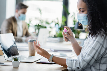 african american businesswoman applying hand sanitizer near colleague working on blurred background