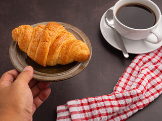 Hand holding of a croissant on a plate with a white coffee cup and a cloth placed on the old kitchen table. Space for text. Vintage background. Concept of dessert, beverage, and relaxation