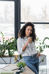 Smiling african american businesswoman holding takeaway drink near papers in office