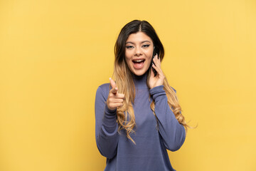 Young Russian girl using mobile phone isolated on yellow background surprised and pointing front