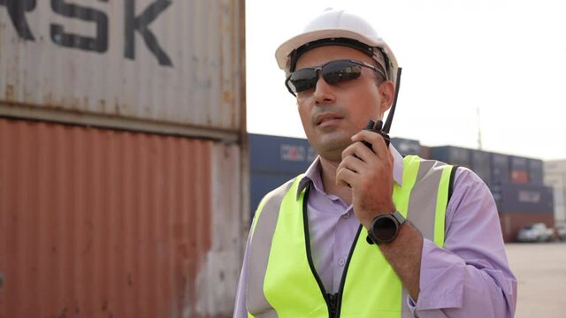 Logistic or engineer foreman talking on radio phone or walkie-talkie with colleague in container warehouse. inspecting for quality control. shipping delivery and export import concept