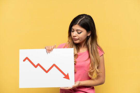 Young Russian girl isolated on yellow background holding a sign with a decreasing statistics arrow symbol with sad expression