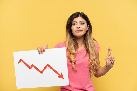 Young Russian girl isolated on yellow background holding a sign with a decreasing statistics arrow symbol with fingers crossing