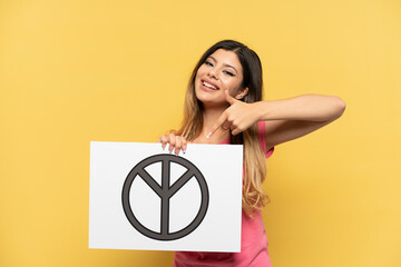Young Russian girl isolated on yellow background holding a placard with peace symbol and  pointing it