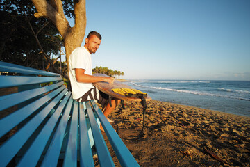 Man surfer sits on a blue bench and waxing yellow surfboard by the ocean.