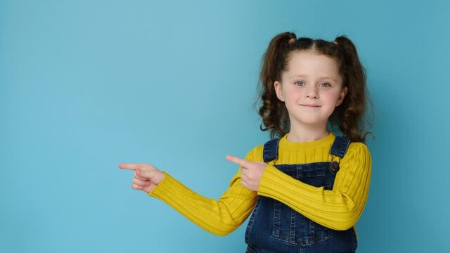 Charming sweet cute little kid girl pointing index fingers aside for advertisement, happy look at camera, standing isolated on blue studio wall background with copy space. Childhood lifestyle concept