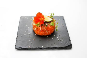 A close-up of a chopped salmon tartare on a black stony tray, on a white background. Appetizer prepared from fresh raw salmon and seasonings, decorated with an edible flower.