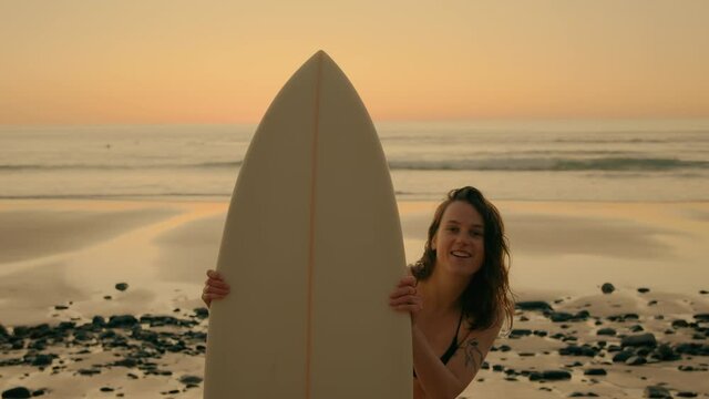 Funny quirky shot of beautiful young woman interact with camera, look from behind surfboard, smile and laugh. Authentic candid portrait of real human with true emotions. Surfing lifestyle vibes