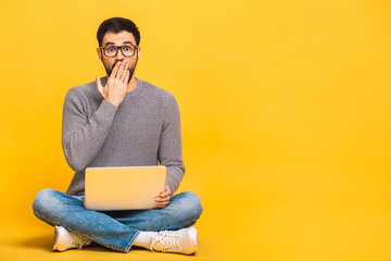 Young bearded man shocked surprised amazed with laptop computer. Funny image of young Caucasian male student model sitting on floor isolated on yellow background.