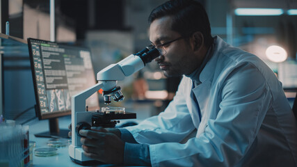Medical Research Scientist Conducts DNA Experiments Under a Digital Microscope in a Biological Applied Science Laboratory. Multiethnic Lab Engineer in Glasses in White Coat Working on Vaccine.