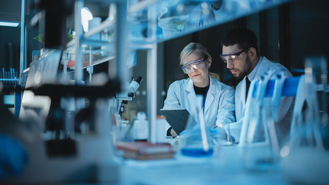 Female and Male Medical Research Scientists Have a Conversation While Conducting Experiments in Sample Test Tubes, Writing Analysis Results on a Tablet Computer. Modern Biological Science Laboratory.