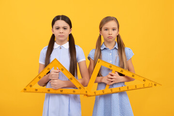 Unhappy school age children hold triangular rulers for geometry lesson yellow background, homework