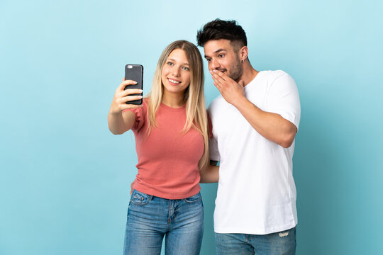Young caucasian couple isolated on blue background making a selfie with the mobile