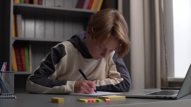 Close-up face of overworked pupil boy doing homework, taking notes in workbook and falling asleep dropping head on desk at home by window. Concept of distance education. Tracking shot in slow motion.